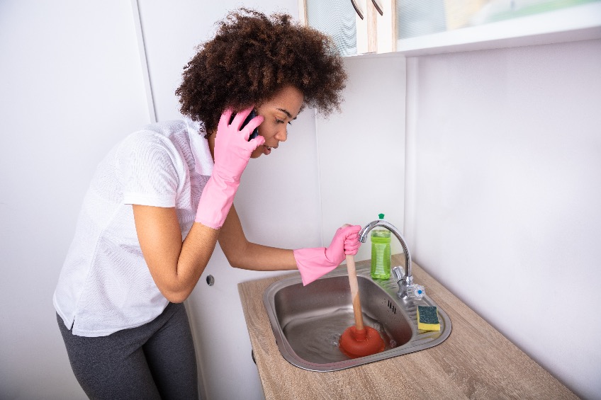Close-up Of A Woman Calling Plumber On Mobile Phone While Cleaning The Sink With Plunger